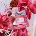 Dior Miss Dior Parfum pour femme, dans un flacon en verre texturé avec un nœud en tissu, se trouve parmi des fleurs et des pétales d'un rose éclatant. Un éclairage doux et chaleureux renforce l'ambiance romantique et élégante. Dakar