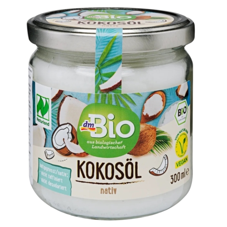 Un pot en verre contenant de l'huile de coco biologique dmBio avec un couvercle argenté, avec des images de noix de coco et de feuilles vertes sur l'étiquette. L'étiquette porte la mention "Kokosöl nativ", ainsi que les logos des certifications végétalienne et biologique. Dakar