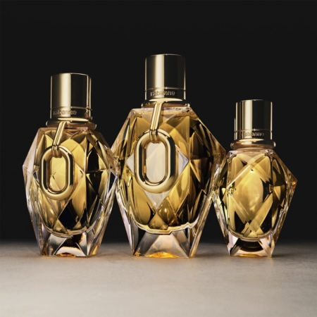 Trois flacons à facettes de l'Eau de Parfum pour Femme Rabanne Million Gold For Her, de tailles différentes, avec des bouchons et des bagues en or métallique, sont présentés sur une surface neutre, sur un fond sombre. Dakar