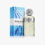 Un flacon de parfum Eau de Rochas, au design en verre texturé et au bouchon argenté, est présenté à côté de sa boîte bleue et blanche, qui comporte des graphiques inspirés de l'eau et le nom de la marque. Dakar