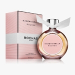 Un flacon de parfum Mademoiselle Rochas avec un ruban blanc sur le bouchon est placé à côté de son coffret rayé rose et blanc, tous deux portant le nom de la marque "Rochas Paris" sur la face avant. Dakar