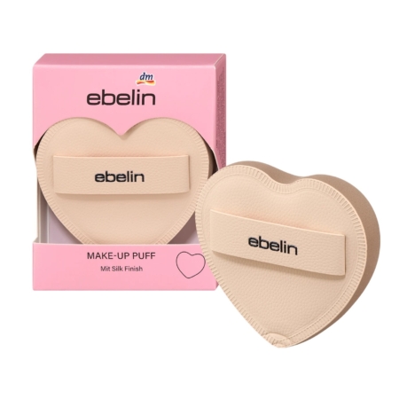 Une houppette de maquillage beige en forme de cœur avec le mot "ebelin" sur sa lanière, présentée à côté de sa boîte d'emballage rose, qui comporte également le logo "ebelin" et des informations sur le produit. Dakar