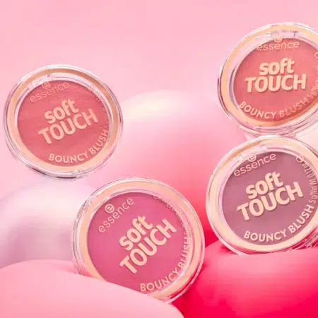 Quatre poudriers Essence Soft Touch Bouncy Blush dans différentes teintes de rose et de pêche sont présentés sur des surfaces arrondies rose pastel et blanches avec un fond rose. Dakar