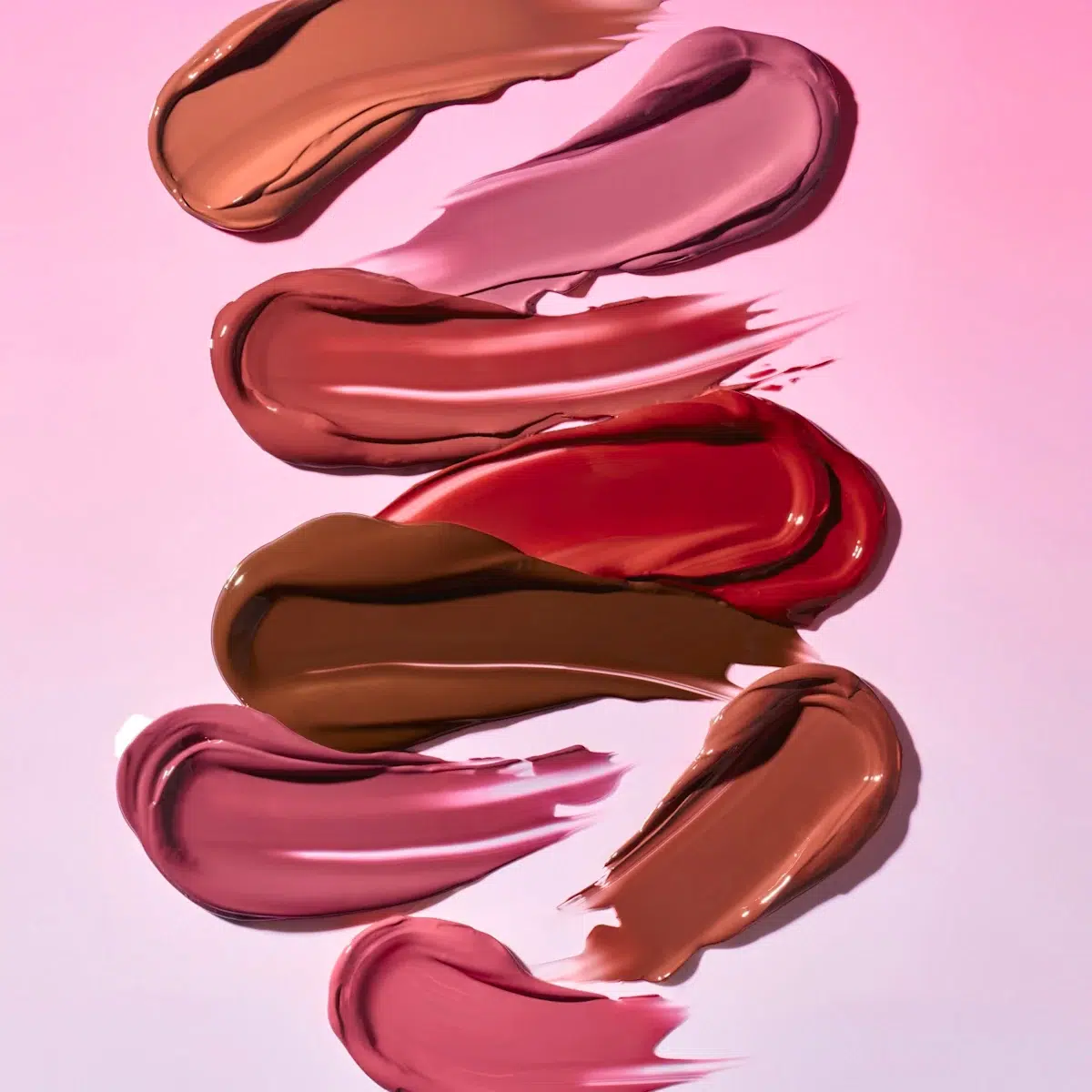 Six échantillons de rouge à lèvres de différentes nuances de rose, de rouge, de mauve et de brun sont étalés artistiquement en zigzag sur un fond rose clair. Dakar