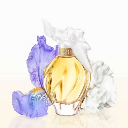 Un flacon de parfum doré avec un bouchon sculpté blanc et orné se trouve entre un iris violet et un œillet blanc, le tout sur un fond clair. Dakar