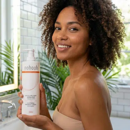 Une femme aux cheveux bouclés sourit en tenant un grand flacon de lotion blanchissante et revitalisante Nohooh d'Univers Cosmetix dans une salle de bain lumineuse avec des plantes et des carreaux blancs en arrière-plan. Dakar