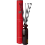 Une haute boîte cylindrique rouge étiquetée "The Ritual of Ayurveda" se trouve à côté d'un flacon diffuseur en verre strié foncé avec plusieurs longs roseaux noirs déployés en éventail au sommet. Dakar