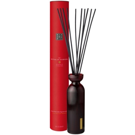 Une haute boîte cylindrique rouge étiquetée "The Ritual of Ayurveda" se trouve à côté d'un flacon diffuseur en verre strié foncé avec plusieurs longs roseaux noirs déployés en éventail au sommet. Dakar