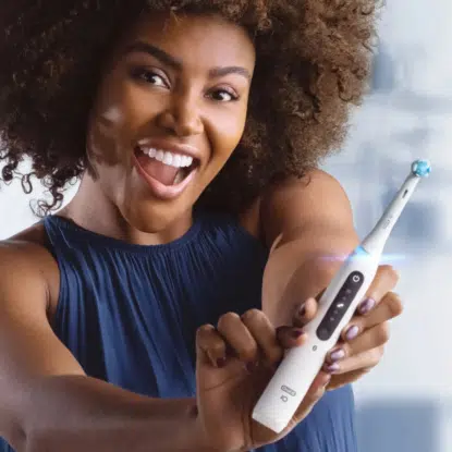 Une femme souriante aux cheveux bouclés tient une brosse à dents électrique Univers Cosmetix et la montre du doigt avec enthousiasme. Elle est vêtue d'un haut bleu marine sans manches dans une pièce très éclairée. Dakar