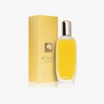 Un flacon de parfum Clinique Aromatics Elixir avec un bouchon doré est placé à côté de sa boîte jaune texturée comportant un emblème floral et le nom du produit. Le fond est blanc. Dakar