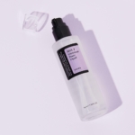 Un flacon en plastique transparent de Cosrx AHA 7 Whitehead Power Liquid 100 ml avec une pompe noire et un bouchon transparent repose sur une surface violette claire. L'étiquette noire et blanche présente de subtils accents violets. Dakar