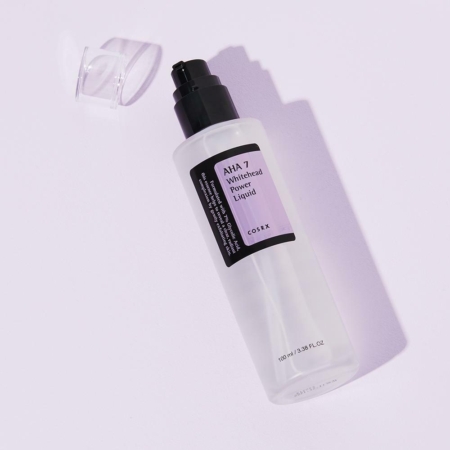 Un flacon en plastique transparent de Cosrx AHA 7 Whitehead Power Liquid 100 ml avec une pompe noire et un bouchon transparent repose sur une surface violette claire. L'étiquette noire et blanche présente de subtils accents violets. Dakar