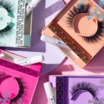 Vue aérienne de trois ensembles de faux cils emballés avec des tubes de colle, placés sur des fonds de couleur pastel (rose, violet et vert menthe). L'emballage comporte un texte sur les boucles et la longueur naturelle. Dakar