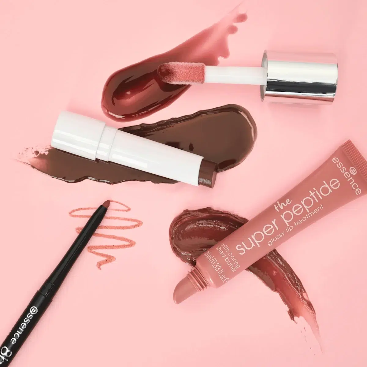 Une présentation à plat de divers produits pour les lèvres sur un fond rose, y compris un brillant à lèvres avec applicateur, un rouge à lèvres, un crayon à lèvres et un tube de "super peptide glossy lip treatment", avec des échantillons de chaque produit. Dakar