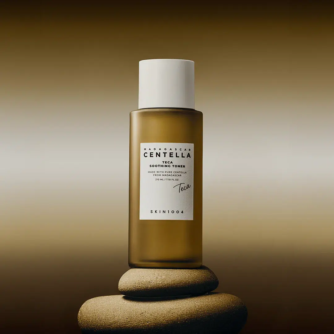 Un flacon brun de SKIN1004 Madagascar Centella Tea-Trica Soothing Toner avec un bouchon blanc est posé sur des pierres lisses empilées sur un fond dégradé de brun doux. Dakar