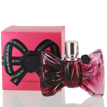 Un flacon de parfum rose en forme de nœud avec un bouchon argenté se trouve devant une boîte rose assortie étiquetée "Bonbon" par Viktor & Rolf. La boîte est ornée d'un nœud noir et d'un texte blanc. Dakar
