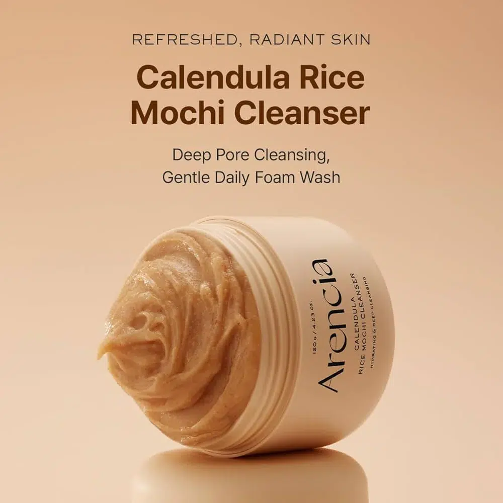 Le pot ouvert d'Arencia Nettoyant Doux Mochi au Riz et au Calendula Peau Sensible et Sèche 120g montre un nettoyant crémeux beige sur un fond légèrement bronzé. Le texte met l'accent sur la peau rafraîchie, le nettoyage en profondeur et la douceur de la mousse de lavage quotidienne. Dakar