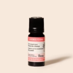 Un petit flacon foncé d'Aroma Zone Actif Pousse des Cheveux 10 ml avec un bouchon noir et une étiquette blanc-rose est représenté sur un fond beige clair. Dakar