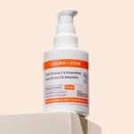 Un flacon d'Aroma Zone Crème Vitamine C et Astaxanthine Bio Éclat du Teint 50ml avec une pompe blanche et orange est posé sur une surface beige sur un fond clair, avec une étiquette indiquant sa certification vegan et biologique. Dakar