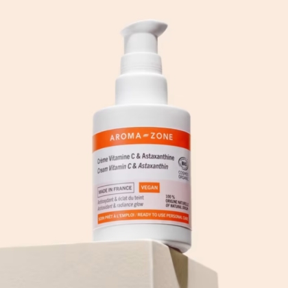 Un flacon d'Aroma Zone Crème Vitamine C et Astaxanthine Bio Éclat du Teint 50ml avec une pompe blanche et orange est posé sur une surface beige sur un fond clair, avec une étiquette indiquant sa certification vegan et biologique. Dakar