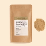 Un sachet en papier marron étiqueté "Aroma Zone Extrait de Plante en Poudre de Kapoor Kachli 100g" avec des motifs floraux et un texte en français est placé à côté d'un petit monticule de poudre marron clair sur un fond beige. Dakar