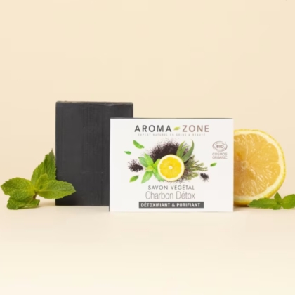 Un savon artisanal noir Charbon Détox Détoxifiant et Purifiant 100g d'Aroma Zone Savon se trouve à côté de sa boîte blanche, avec des images de charbon de bois, de menthe et de citron. Des feuilles de menthe fraîche et un demi-citron sont posés à côté du savon sur une surface claire. Dakar