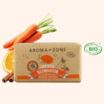 Une barre Aroma Zone Savon Végétal Illuminateur aux Huiles Essentielles 100g est posée au milieu de carottes, d'une rondelle d'orange et de bâtons de cannelle. L'emballage est décoré d'illustrations et affiche le logo de certification des cosmétiques biologiques BIO. Dakar