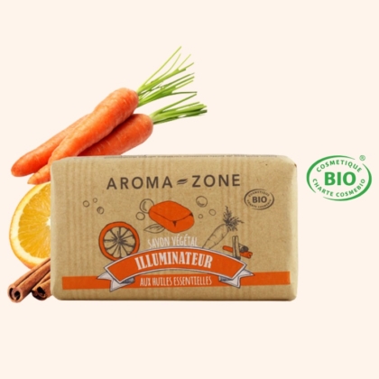 Une barre Aroma Zone Savon Végétal Illuminateur aux Huiles Essentielles 100g est posée au milieu de carottes, d'une rondelle d'orange et de bâtons de cannelle. L'emballage est décoré d'illustrations et affiche le logo de certification des cosmétiques biologiques BIO. Dakar