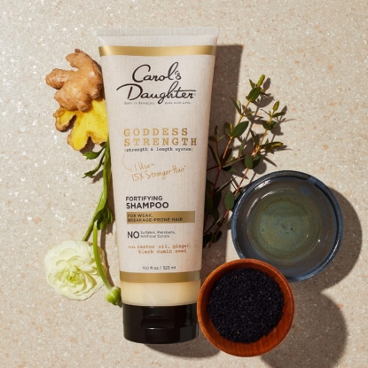 Un tube de shampooing fortifiant Carol's Daughter Goddess Strength d'Univers Cosmetix est entouré de gingembre, d'une fleur jaune, d'une fleur blanche, d'un plat en céramique bleue contenant un liquide et d'un petit bol noir avec des graines noires sur une surface mouchetée. Dakar