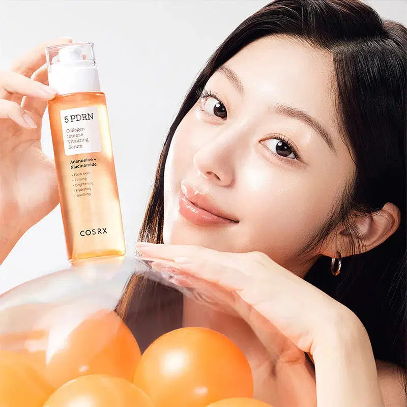 Une femme aux longs cheveux bruns brandit un flacon de COSRX 5 PDRN Collagen Serum et sourit. Elle est entourée de ballons orange et son menton repose délicatement sur l'un d'entre eux. Dakar