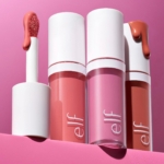 Trois tubes de rouge à lèvres liquide e.l.f. dans les tons pêche, rose et nude se tiennent debout sur une surface rose ; un applicateur présente une teinte pêche crémeuse - idéale pour l'associer au Blush Elf Fard à Joues Liquide Camo Ultra-Pigmenté et Longue Tenue 4ml. Dakar