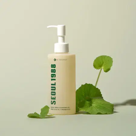 Un flacon pompe beige de KSecret Huile Nettoyante Séoul 1988 Cica de Pin 1 % + Probiotiques 200 ml se tient debout sur une surface légère entourée de feuilles vertes. Le design minimaliste présente un texte épuré et une ombre douce en dessous. Dakar