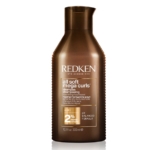 Un flacon brun du shampooing Redken All Soft Mega Curls, avec un texte et des graphiques blancs et dorés, mettant en valeur des ingrédients tels que l'aloès et un complexe nourrissant à 2 %, d'une contenance de 300 ml. Dakar