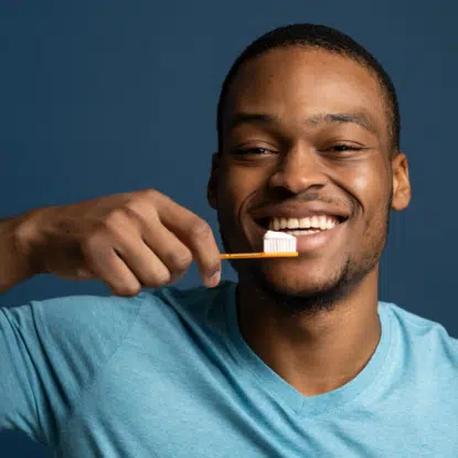 Un homme souriant, vêtu d'un t-shirt bleu clair, tient une brosse à dents près de ses dents, posant comme s'il se brossait, sur un fond bleu foncé - montrant une confiance éclatante avec Univers Cosmetix. Dakar