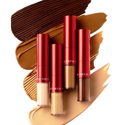 Quatre tubes de Correcteur Floutant Double Action Glide an Hide avec des bouchons rouges métalliques sont disposés verticalement au-dessus d'échantillons de fond de teint dans différentes teintes de beige clair à foncé et de brun, sur un fond blanc. Dakar