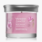 Une bougie Yankee rose étiquetée "Wild Orchid" dans un pot en verre transparent avec un couvercle argenté, avec des illustrations de fleurs d'orchidées et pesant 122g. Dakar