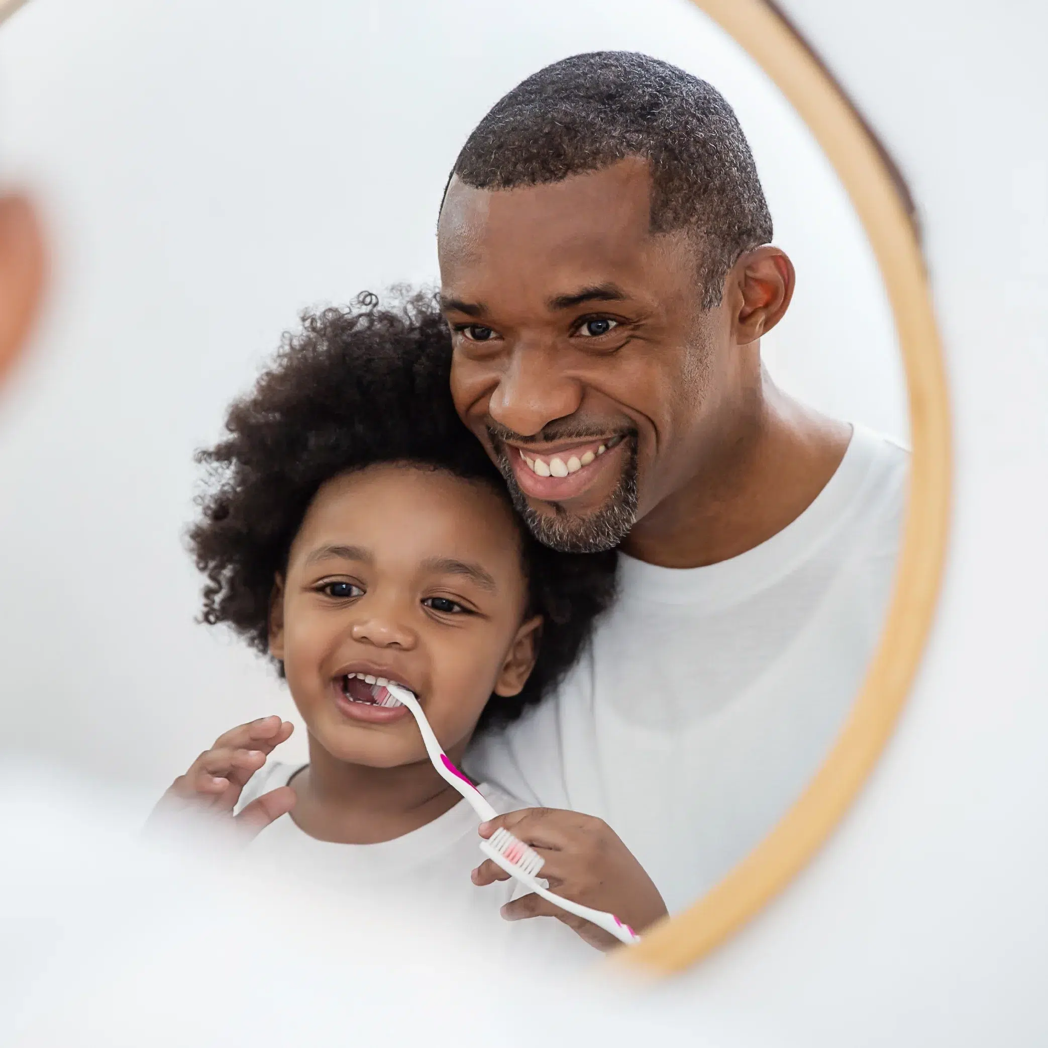 bucco dentaire Un homme souriant aide un jeune enfant à se brosser les dents, tous deux se reflétant dans un miroir rond de salle de bains. L'enfant, qui utilise une brosse à dents Univers Cosmetix, semble heureux tandis que l'homme le guide doucement. Dakar