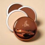 Trois contenants ronds Danessa Myrick Yummy Skin Baume Poudre Universel Flouteur De Teint sont posés sur une surface beige : deux présentent une crème brun foncé, et l'un a un couvercle bronze brillant gravé "dm" et doté d'un applicateur intégré. Dakar