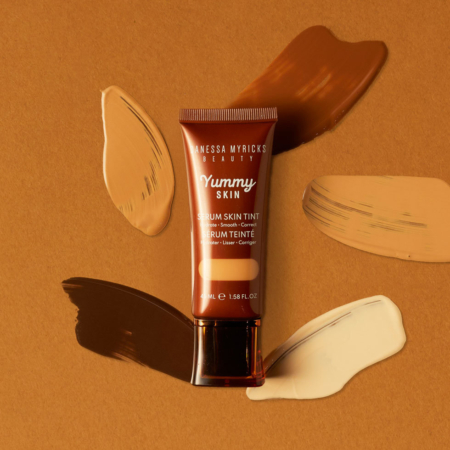 Un tube de Danessa Myricks Beauté Fond de Teint Teinté Yummy Skin Sérum aux Peptides et Céramides 45ml est posé sur une surface brune, entouré d'échantillons de fond de teint dans des teintes beige, marron, beige et crème. Dakar