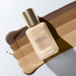 Un flacon de fond de teint Estée Lauder Double Wear Stay-in-Place Makeup repose sur un arrière-plan présentant des échantillons de fond de teint dans différentes nuances de brun et de beige, représentant différents teints de peau. Dakar