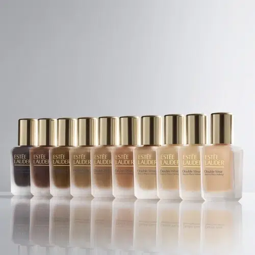 Dix flacons de fond de teint Estée Lauder Double Wear sont disposés en rangée, présentant un dégradé de teintes allant du foncé au clair, avec des bouchons dorés et un arrière-plan minimaliste. Dakar