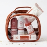 Une trousse de toilette transparente avec une bordure marron contient plusieurs produits de beauté de la marque MUSC, notamment des lotions, des sprays, des gels douche, des lingettes pour le visage et des petites boîtes, le tout avec un emballage blanc et rose-rouge assorti. Dakar