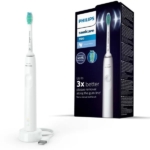 Une brosse à dents électrique Philips Sonicare 3100 se tient debout sur sa base de chargement, à côté de sa boîte, sur laquelle figurent l'image de la brosse à dents, sa marque et des informations sur ses capacités d'élimination de la plaque dentaire. Dakar