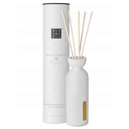 Un flacon diffuseur en céramique blanche avec plusieurs bâtonnets de roseau à côté d'une grande boîte cylindrique étiquetée "Rituals... The Ritual of Sakura" et "blossoming blossoms fragrance oil, rice milk & cherry blossom. Dakar