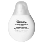 Le baume à lèvres Squalane + Acides Aminés de The Ordinary se présente sous la forme d'un tube blanc et arrondi, avec un texte noir et un bouchon transparent. L'emballage affiche les détails du produit en anglais et en français. Dakar