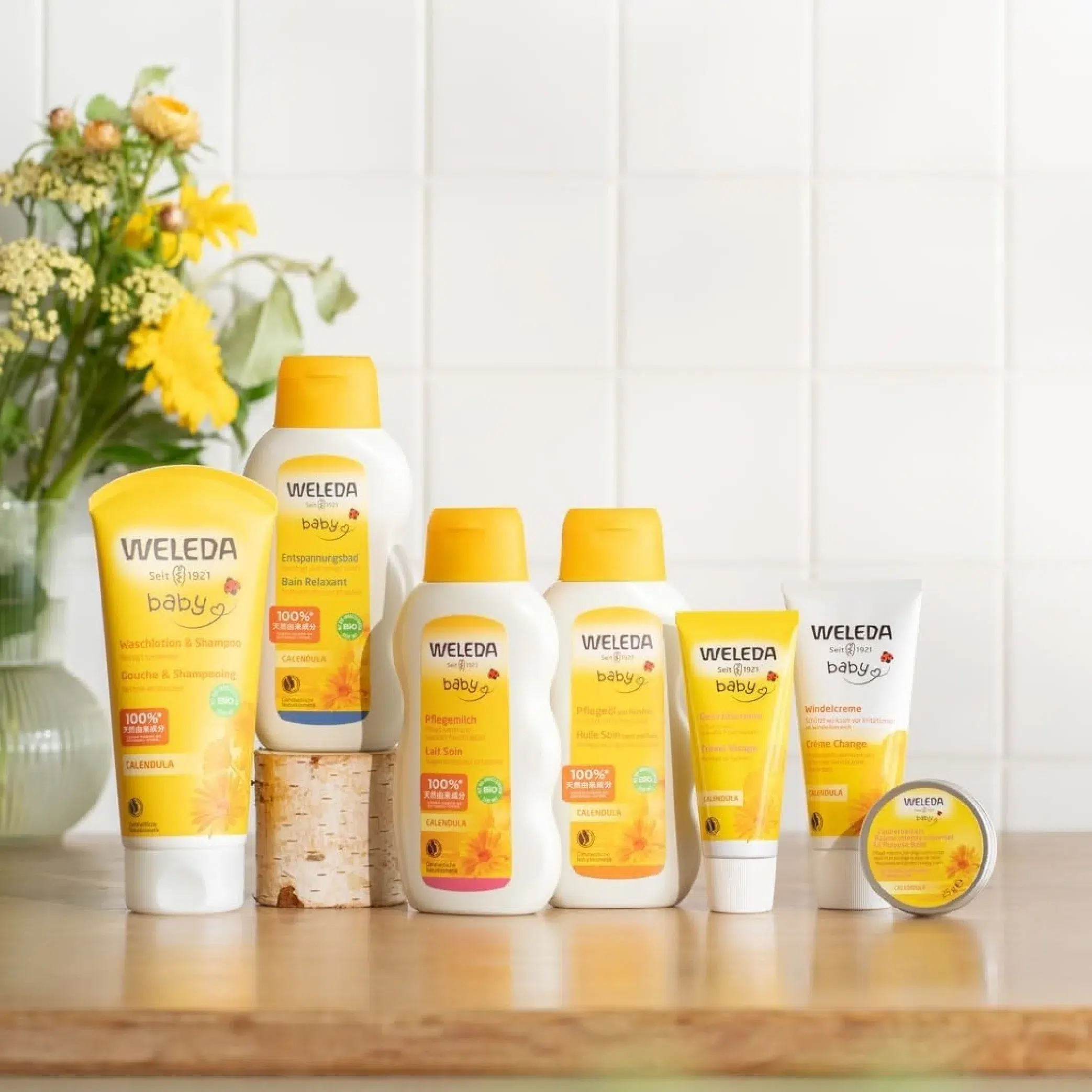 weleda baby Une collection de produits de soins pour bébés Weleda d'Univers Cosmetix, dont l'emballage est jaune et blanc, est disposée sur une surface en bois, complétée par des fleurs jaunes et un mur carrelé blanc en arrière-plan. Dakar