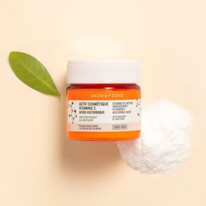 Un pot d'Aroma Zone Actif Acide Ascorbique Vitamine C Pure Éclat du Teint, de 10 g, repose sur une surface beige, à côté d'une feuille verte et d'une poudre blanche ; son étiquette comporte des mentions en français et en anglais. Dakar