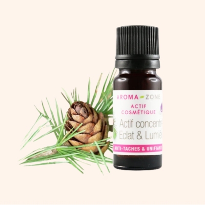 Un petit flacon marron d'Aroma Zone Actif Concentré Éclat et Lumière Anti-Taches Unifiant, d'une contenance de 30 ml, est posé à côté d'une branche de pin et d'une pomme de pin sur un fond beige clair. Dakar
