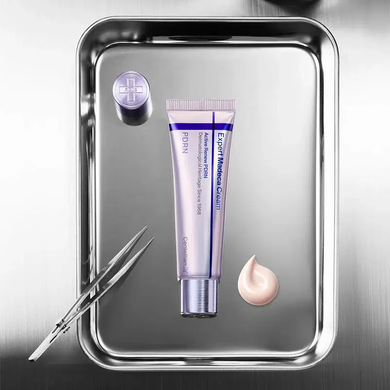 Un plateau métallique contient un tube de Dr Althea Reju 5000 Crème Hydratante Apaisante 20ml (Copie), une petite cuillère de crème, une pince à épiler et un bouchon rond sur une surface en acier inoxydable. Dakar