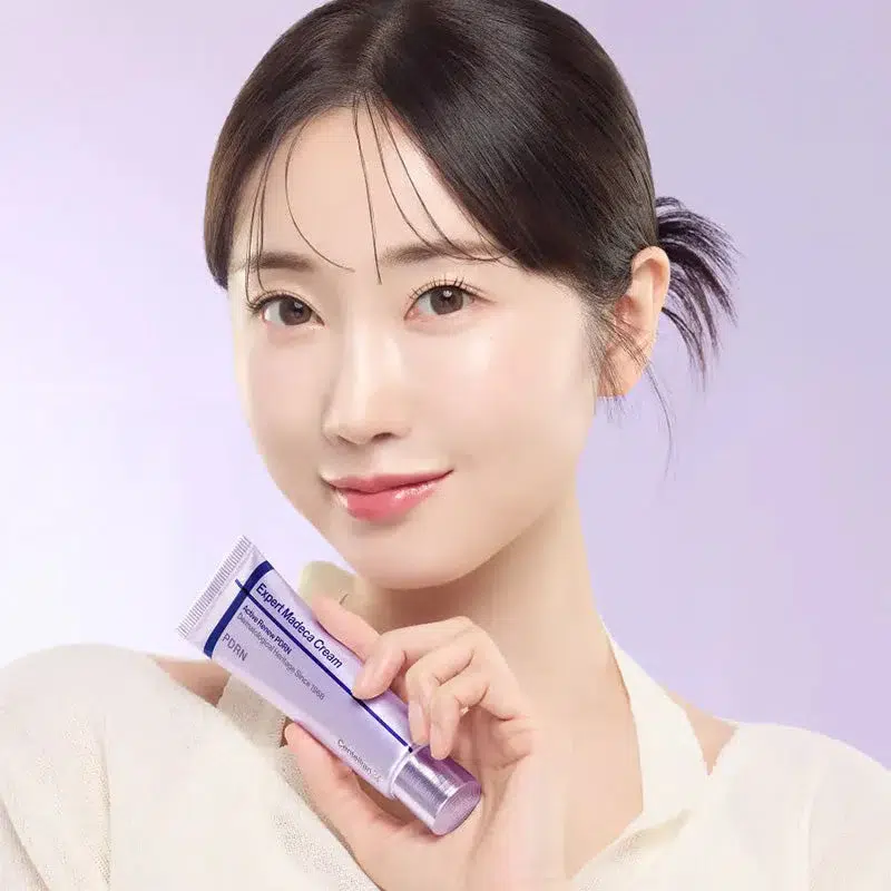 Une femme à la peau claire et aux cheveux foncés sourit doucement, tenant un tube lavande de Dr Althea Reju 5000 Crème Hydratante Apaisante 20ml (Copie) sur un fond violet clair. Dakar